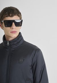 Giacca nera imbottita con colletto alto e chiusura con zip, caratterizzata da una patch con logo. Occhiali da sole neri oversize con un design geometrico.