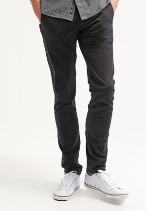 Man draagt slim-fit zwarte broek, witte sneakers en een grijs patroonoverhemd met handen in de zakken, staand tegen een witte achtergrond.
