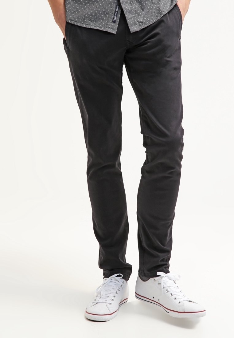 Homme portant un pantalon noir slim, des baskets blanches et une chemise grise à motifs, les mains dans les poches, debout devant un fond blanc.