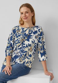 Blouse florale avec une base crème ornée de motifs de fleurs bleues et brunes, manches 3/4 et coupe décontractée. Tissu lisse et texturé.