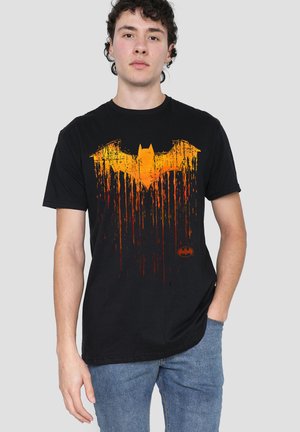 Young man wearing black t-shirt with large distressed orange Batman logo, blue jeans, standing against plain light gray background.