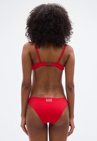 Σετ κόκκινου μπικίνι με λεπτές τιράντες, κλείσιμο πίσω με αγκράφα και λογότυπο στη μέση. Ομαλή υφή και μινιμαλιστικός σχεδιασμός.