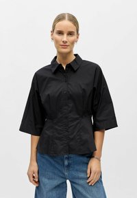 Femme aux cheveux blonds portant une chemise noire boutonnée à manches trois-quarts et un jean bleu, debout devant un fond clair uni.