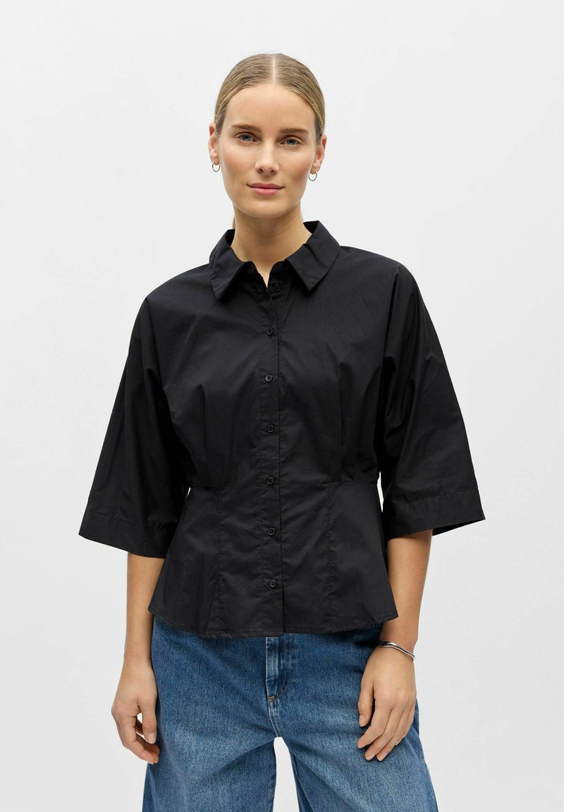 Femme aux cheveux blonds portant une chemise noire boutonnée à manches trois-quarts et un jean bleu, debout devant un fond clair uni.