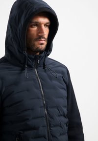 Marineblauwe pufferjack met capuchon, voorzien van een ritssluiting aan de voorkant, elastische accenten en gestructureerde schouderpanelen, ontworpen voor warmte en comfort.