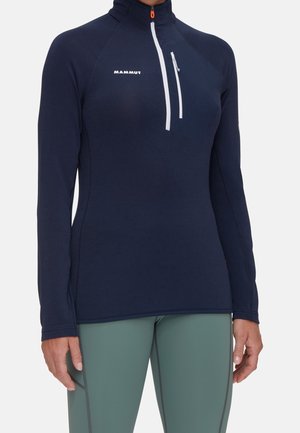 Persoon die een marineblauw topje met lange mouwen en ritssluiting met het "Mammut" logo en lichtgroene aansluitende broek draagt.