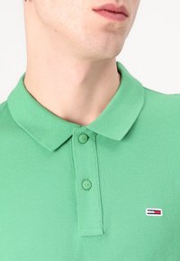 Grønn polo skjorte laget av teksturert stoff, med krage og to knapper. Merkelig logo med røde, hvite og blå striper på brystet.