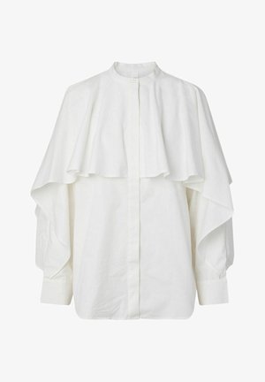 Valge pikkade varrukatega pluus kõrge kraega ja kihtidena kaela ning ülakeha kattev kapuutsilaadne kangas, kaelas nööbitud.