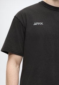 Κοντινό πλάνο ενός ατόμου που φοράει μαύρο t-shirt με τη μάρκα ARKK, δείχνοντας τον άνω κορμό και το αριστερό χέρι σε απλό φόντο.