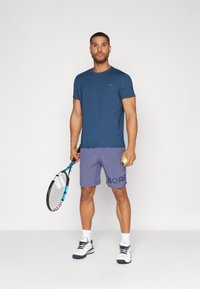 Krátky rukáv námornícky modrý tričko, purpurové šortky s textom "BORGO", drží tenisovú raketu a loptičku, obuté biele a námornícky modré športové topánky.