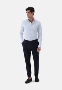 Helleblaues Button-Down-Hemd, marineblaue Hose und dunkle Loafers. Das Hemd hat einen Standardkragen und lange Ärmel; glatte Textur.