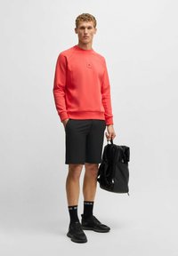 Felpa corallo con maniche raglan, pantaloni corti neri e sneakers nere. Il modello tiene uno zaino nero con spallacci regolabili.