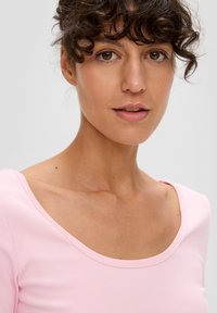 Jasnoróżowa, prążkowana bluzka z szerokim dekoltem i krótkimi rękawami. Gładka tekstura z subtelnymi wariacjami kolorystycznymi. Zbliżenie na górną część tułowia.