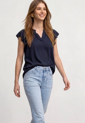 Jeune femme aux longs cheveux bruns portant un haut bleu marine à manches courtes, un jean bleu clair et des boucles d'oreilles dorées en forme de fleur, souriante et marchant vers l'avant.