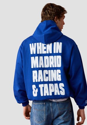 Hombre con sudadera azul con capucha con gran texto blanco en la espalda que dice «"WHEN IN MADRID RACING & TAPAS"» sobre fondo liso.