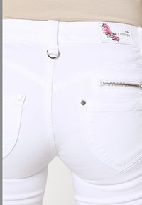 Los pantalones vaqueros de mezclilla blanca presentan un diseño ajustado con bolsillos traseros, herrajes plateados, una etiqueta floral y un bolsillo con cremallera en el lateral.
