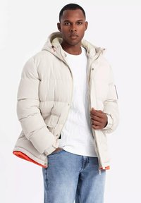 Beige pufferjacka med huva, med dragkedja fram, elastiska ärmslut och orange foder. Bärs över en vit mönstrad sweatshirt och blå jeans.