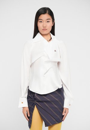 White structured shirt with long sleeves, featuring a twisted front design and a small logo. Paired with a navy striped skirt and yellow tights.