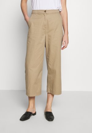 Osoba ubrana w beżowe szerokie spodnie o kroju cropped z guzikiem i suwakiem, w połączeniu z czarnymi butami wsuwanymi i białą bluzką.