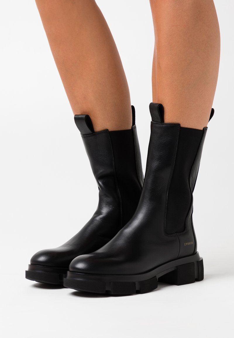 Bottes en cuir noires mi-mollet avec semelles épaisses portées sur des jambes nues sur fond blanc.