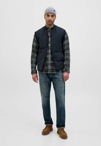 Gilet trapuntato blu navy sopra una camicia a quadri verde, abbinato a jeans azzurri chiari e scarpe in stile mocassino marroni. Sovrapposizione casual e contrasto di texture.