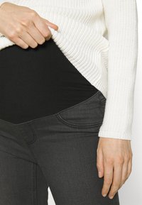 Maglione bianco a costine indossato sopra a un body nero, abbinato a jeans grigio scuro con tasche curve e una texture liscia.