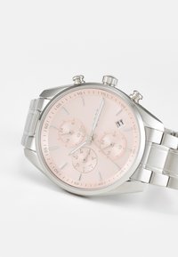 Srebrna ura iz nerjavnega jekla s psih dialom, tremi subdiali, prikazom datuma in povezanim zapestjem. Ima minimalistične oznake ur.