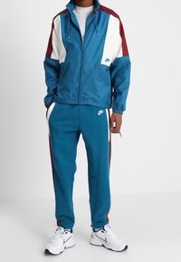 Ensemble de survêtement Nike pour homme comprenant une veste bleue avec des accents blancs et bordeaux, fermeture éclair, et un pantalon bleu assorti avec des bandes latérales et le logo.