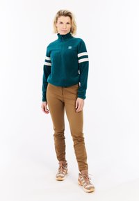 Golf pullover in pile color teal con mezza zip, righe bianche sulle maniche, abbinato a pantaloni slim-fit marroni e scarpe da trekking beige con dettagli chiari.