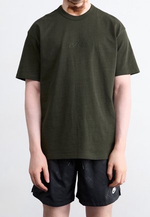 Oliivinvihreä lyhythihainen t-paita, jossa on hienovaraisesti kirjailtu "Nike"-logo, yhdistettynä mustiin shortseihin, joissa on geometrisia kuvioita ja valkoinen logo.