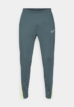 Mørk teal sportsbukser med elastisk talje, tapered ben og klare gule detaljer på siderne. Har et hvidt logo på forsiden.