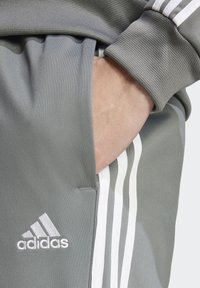 Šedé tepláky Adidas majú štruktúrovanú tkaninu, bočné vrecká a biele tri prúžky pozdĺž nohavíc so bielym logom na prednej strane.