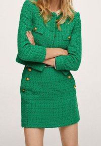 Giacca in tweed verde con bottoni dorati e due tasche sul petto, abbinata a una gonna aderente coordinata. Tessuto testurizzato con un motivo a scacchi.