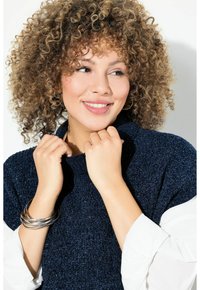 Maglione a maglia testurizzata di un blu navy con colletto alto. Il modello tiene il colletto con entrambe le mani; indossa braccialetti d'argento e una manica bianca.