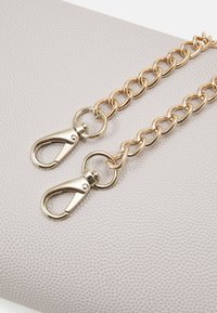 Cintura in metallo dorato con due ganci a clip, appoggiata su uno sfondo grigio chiaro e strutturato. La catena presenta maglie ovali uniformemente distanziate.