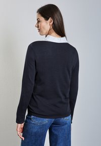 Maglione blu navy scuro con collo rotondo e maniche lunghe, caratterizzato da una camicia bianca con colletto indossata sotto e una trama liscia. Abbinato a jeans in denim blu.