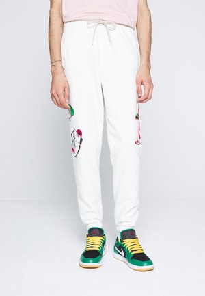 Person iført hvide joggingbukser med farverige lapper og grønne, sorte og gule sneakers, der står mod en ensfarvet baggrund.