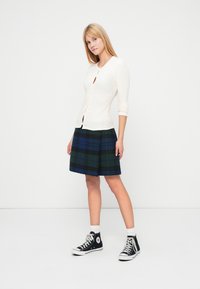 Cardigan crem cu dungi, decolteu rotund și închidere cu nasturi, asortat cu o fustă plisată în carouri în culori închise. Pantofi sport negri înalți completează ținuta.