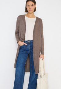 Bruine lange cardigan van zacht materiaal, met een knoopsluiting aan de voorkant en geribbelde manchetten, gecombineerd met een licht top en blauwe flare jeans. Witte tote bag.