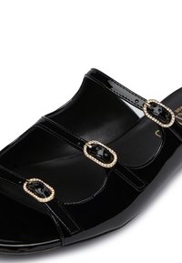 Sandalo in pelle verniciata nera con tre cinturini e fibbie in tonalità oro decorate con strass trasparenti. Finitura liscia e lucida con una suola sagomata.