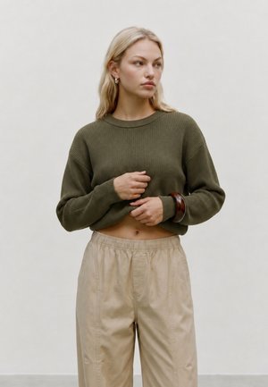 Donna con capelli biondi che indossa un maglione verde oliva e pantaloni beige, che guarda di lato contro uno sfondo chiaro e uniforme.