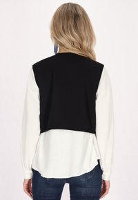 Schwarze verkürzte Weste über einer weißen Langarmbluse. Die Bluse hat einen runden Saum und das Model trägt blaue Ohrringe. Strukturiert sichtbares Material.