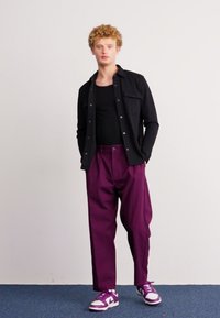Camisa preta de botão sobre um top de alças preto, combinada com calças largas de cintura alta roxas e ténis roxos e brancos.