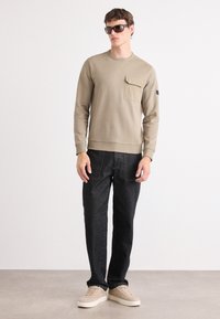 Sweatshirt bege claro com um bolso no peito, combinado com calças de ganga escuras e ténis bege. Modelo a usar óculos de sol escuros. Fundo simples.