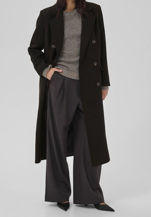 Femme portant un long manteau noir à double boutonnage sur un pull gris, un pantalon large gris foncé et des chaussures noires pointues, debout avec une main dans la poche.