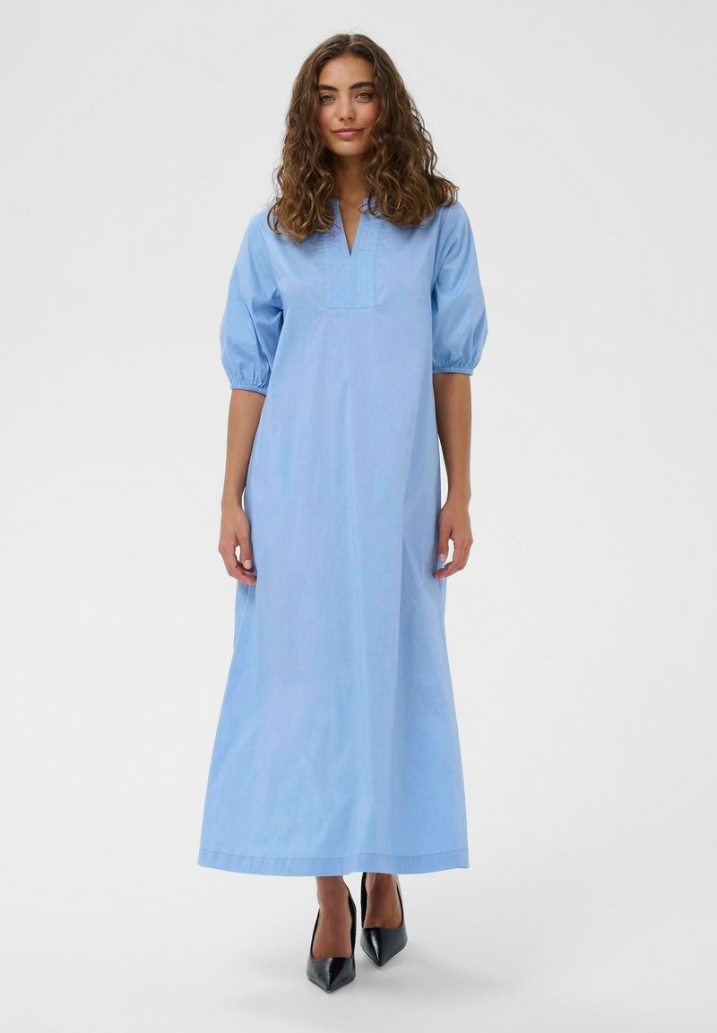 Žena s kovrčavom kosom u svijetloplavoj haljini dugu do gležnjeva s nabranim rukavima i crnim visokim štiklama sa špicom, stoji ispred jednobojne pozadine.