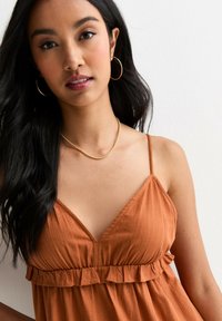 Robe orange rouille avec un ourlet à volants et des bretelles spaghetti ajustables ; tissu doux et texturé avec un décolleté en V. Collier en or et boucles d'oreilles créoles.