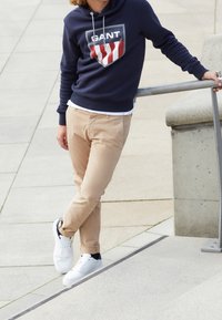 Sudadera con capucha azul marino con logo, pantalones beige claro y zapatillas blancas. Con cordones en la capucha. De pie sobre una superficie pavimentada.