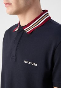 Homme portant un polo bleu marine avec un col rayé rouge, beige et blanc, et le logo « HILFIGER » sur la poitrine.