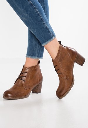 Bottines en cuir marron à lacets et talons carrés portées avec un jean bleu à ourlets effilochés sur fond blanc.